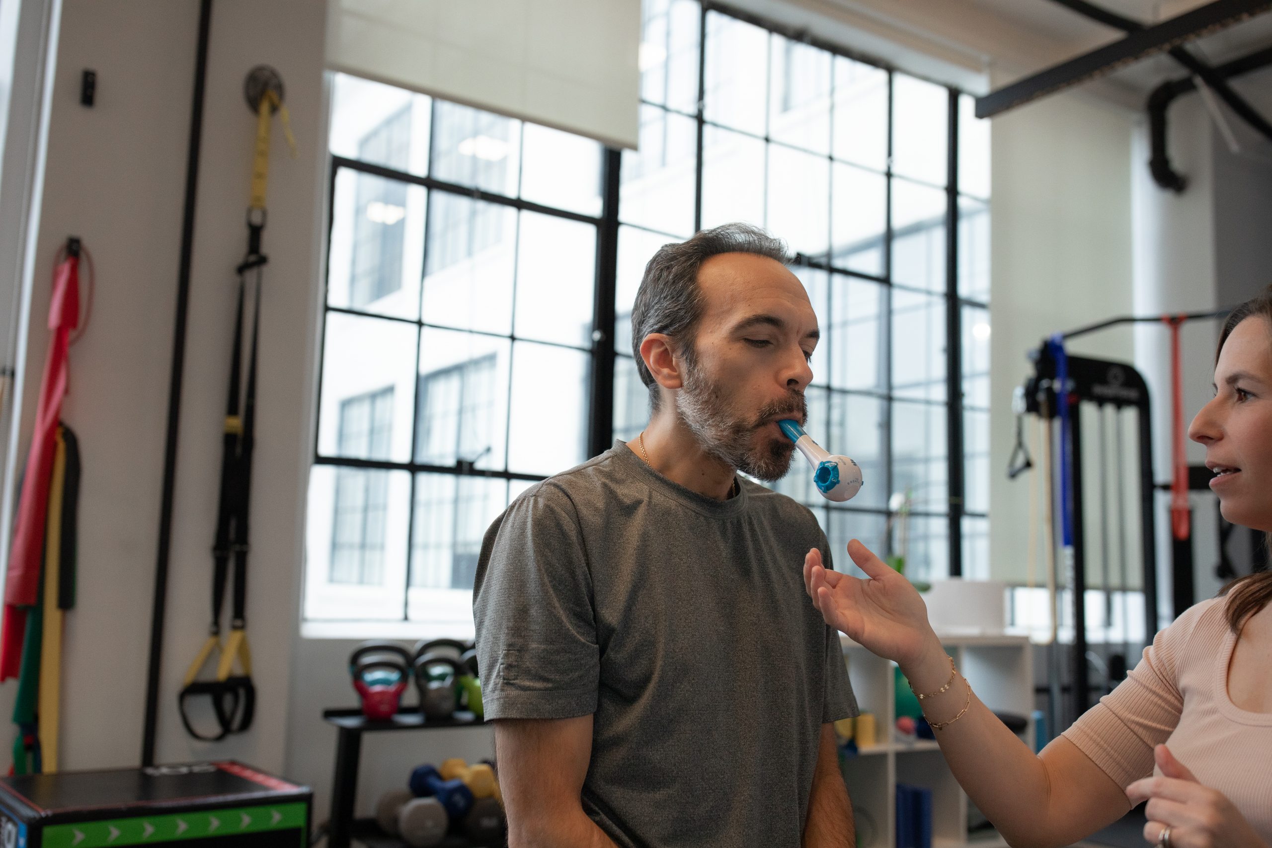 Man in a physical therapy center using a medical device in his mouth. Female physical therapist is supporting the treatment by closely standing by